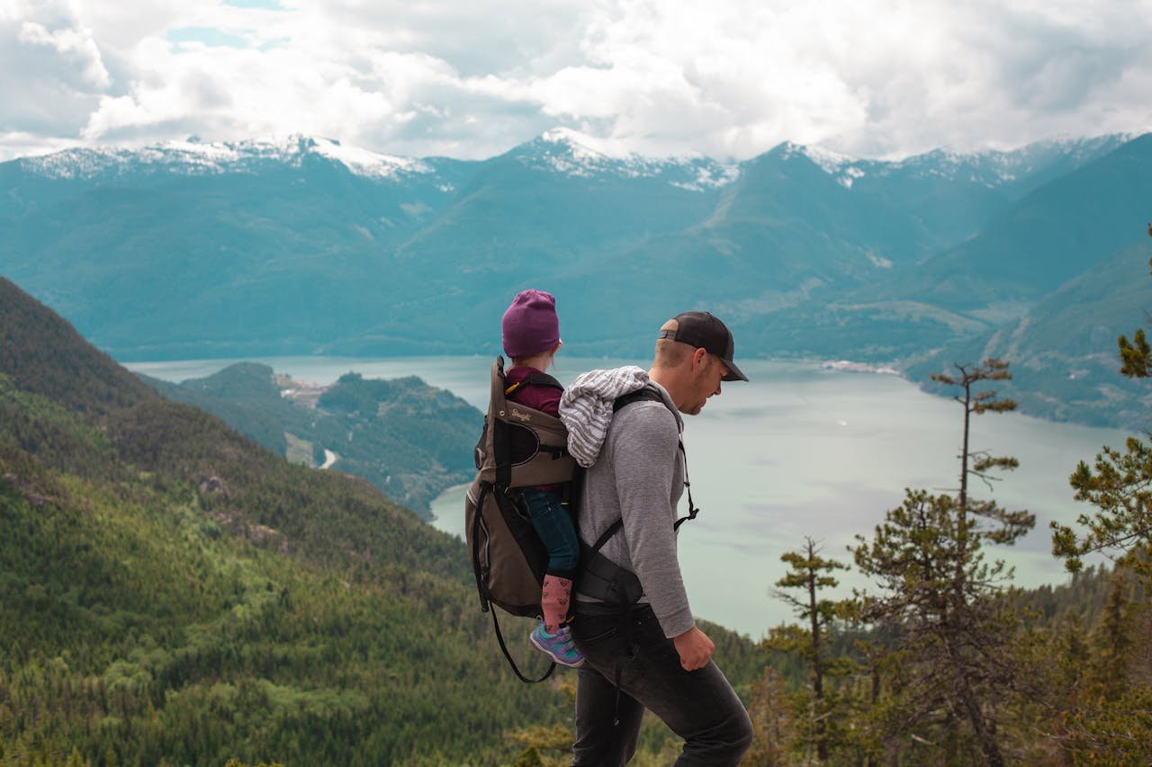 A father carries his child on a scenic mountain trail, embracing adventure and parenthood.