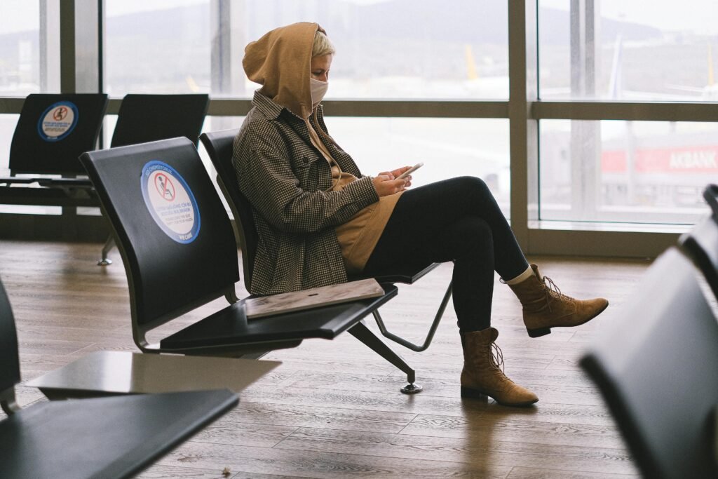 A woman in a hoodie sits in an airport lounge, practicing social distancing while using her phone.